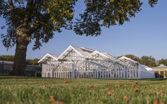 An exterior view of the steel and glass structure of the West Conservatory with a grassy lawn and green tree in the forefront. 