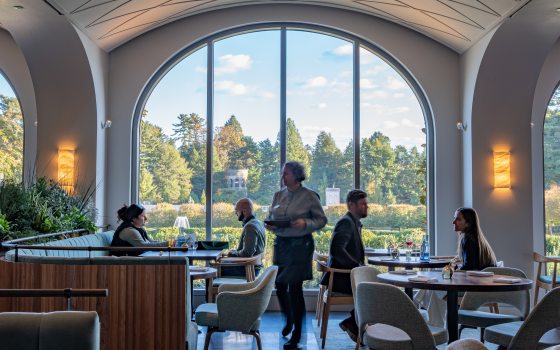 View of a diners seated in a restaurant in front of a large arched window that looks out onto fountains and colorful autumn trees in the distance. 