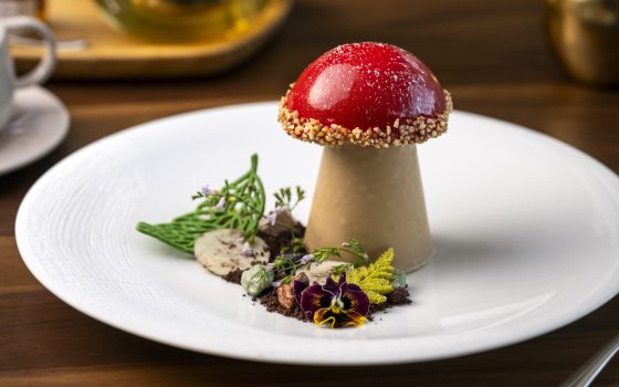 A dessert shaped like a mushroom is dressed with a side of cocoa soil and flowers.