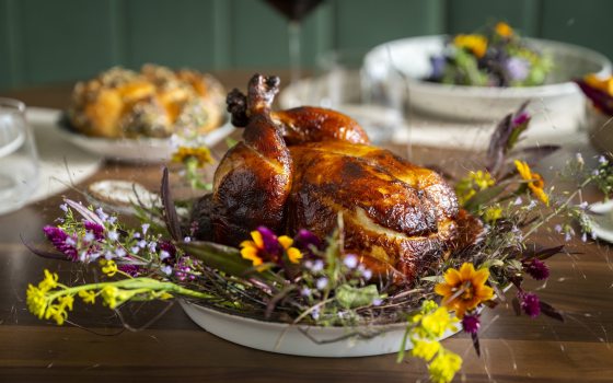 A whole roasted chicken is served in a small casserole skillet on a bed of herbs and flowers.