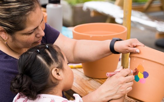 An adult and child paint a multi-colored flower on a large clay pot.