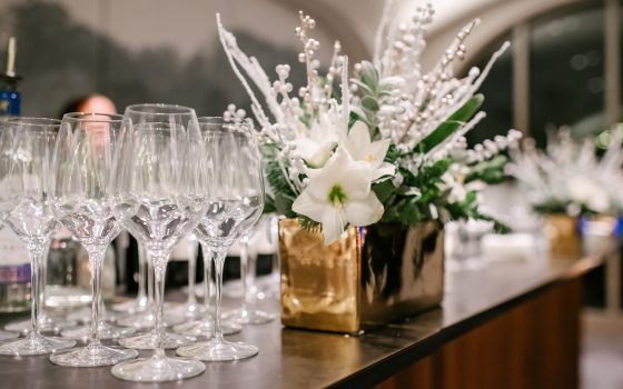 Closeup of array of empty wineglasses on a counter next to a floral arrangement of white blooms and berries in a gold rectangular container.