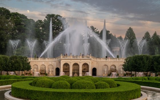 Fountain jets soar high above a fountain with a circular section of green boxwood hedges in front of it.