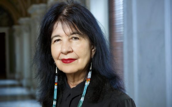 A woman with long black hair, red lipstick, and long earrings looks at the camera. 