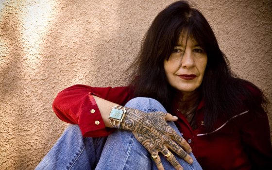 Women in red shirt and jeans sits facing the camera and displaying an intricate tattoo on her hand and a turquoise cuff bracelet 