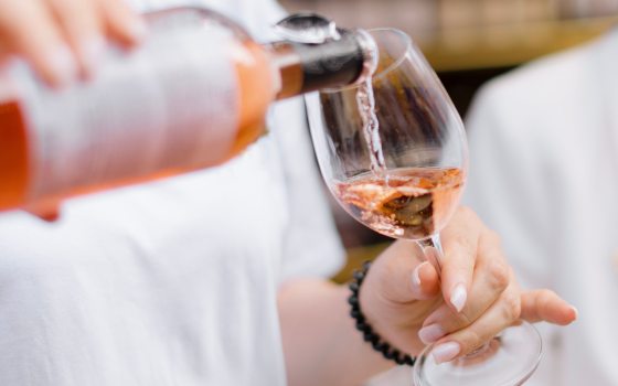 A close-up of a person pouring rosé wine into a clear wine glass at an outdoor event. The wine has a pink hue, and the person holding the glass has a light-colored shirt and manicured nails. The background is softly blurred, emphasizing the action of pouring.