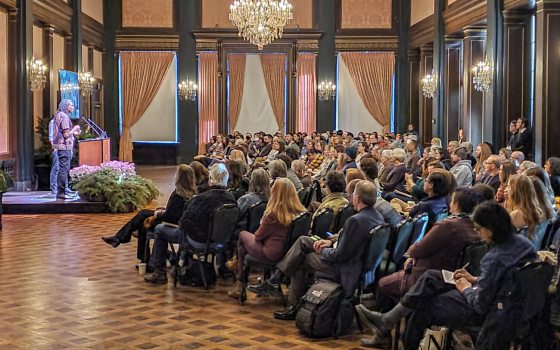 Rows of people in a ballroom listening to a speaker at a podium.