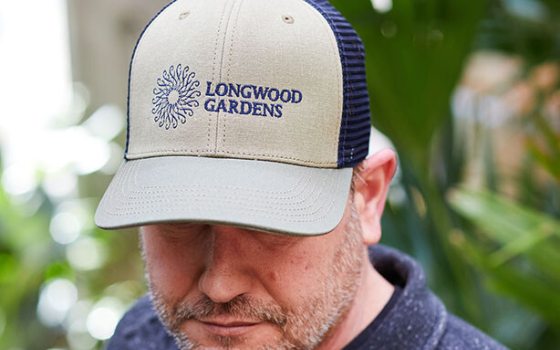 A person modeling a blue and ivory trucker hat with the Longwood Gardens logo on the front. 