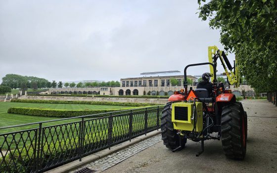 A person in a red tractor trimming trees along an allee with a large yellow shearing device on the front of the tractor.