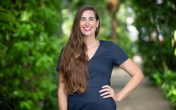 A woman with brown hair wearing a navy dress smiles for the camera while being surrounded by greenery. 