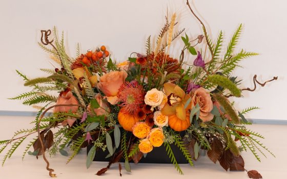 A fall themed centerpiece with flowers in ranges of yellow and orange set against a white backdrop.