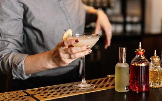 A person's hands behind a bar holding a cocktail glass preparing to hand it to someone. 