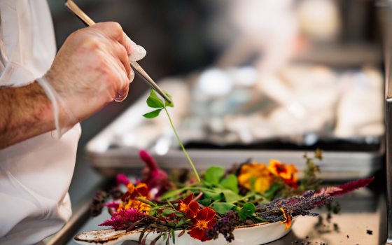 A chef adds a three-leaf clover to a ceramic serving dish filled with green leaves and colorful flowers.