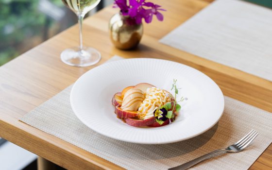 Sliced peaches and cream, drizzled with caramel and topped off with a purple-yellow pansy, fill the center of a shallow white dish.