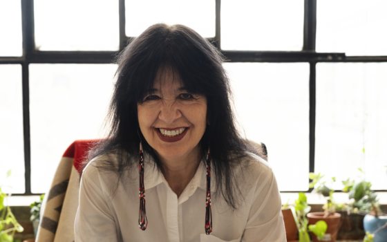 A person in a white shirt, black hair, with long beaded earring smiling at the camera.