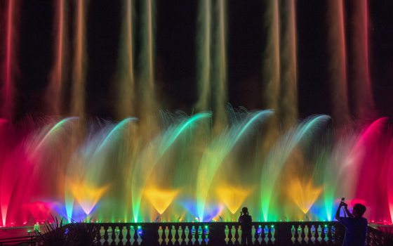 A row of parallel fountain pairs in rainbow colors shoot toward the sky, with a middle layer of dancing fountain spray, and a lower level of cone-shaped fountains.
