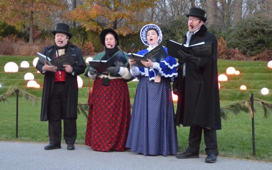 Four singers dressed in old-fashioned Dickens-era clothing and holding black songbooks, stand and sing in front of a lawn decorated with globes of white light.