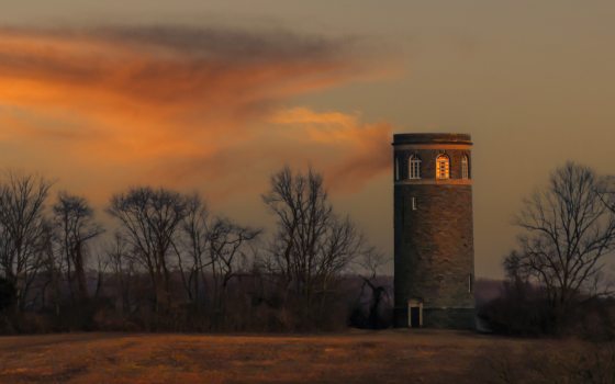 Clouds reflect an orange sunset over a field in winter, with leafless trees in the background and a stone tower on the right.