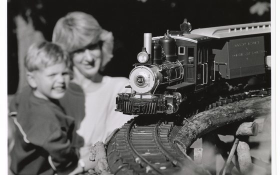 A black and white photograph of a toy or model steam train engine and car running on a curved track in the foreground. In the soft-focus background, an adult woman and a young boy are smiling as they watch the train.