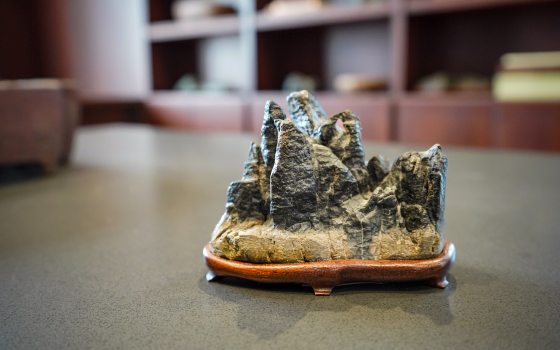  The alt text for this image should be:  A close-up, low-angle shot of a small, dark, craggy rock formation carving, resembling miniature mountains or peaks. It sits on a smooth, dark wooden stand with curved feet. The object is centered on a grey, textured table, with a blurred background of dark wooden shelving or cabinetry.