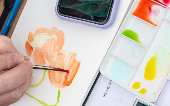 Close-up view of an artist painting an orange watercolor flower on paper, with a palette of vibrant paints and a smartphone visible nearby.