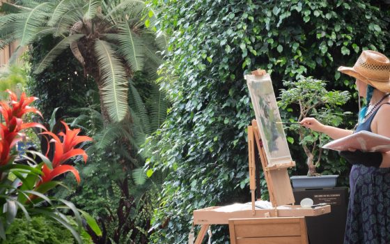 An artist wearing a straw hat paints a bonsai tree on an easel while surrounded by lush tropical plants and red bromeliads in a conservatory.