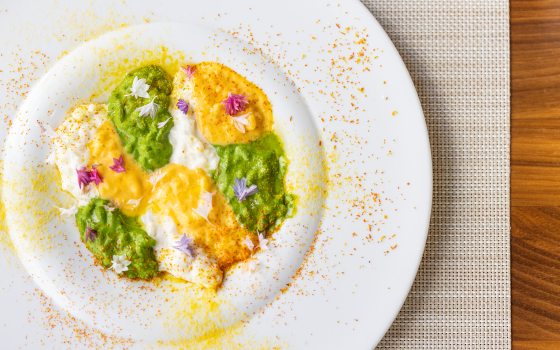 Close-up, overhead view of a plated dish, likely risotto, with alternating dollops of creamy white, vibrant green, and yellow puree or sauce, garnished with edible flowers and surrounded by spice dust.