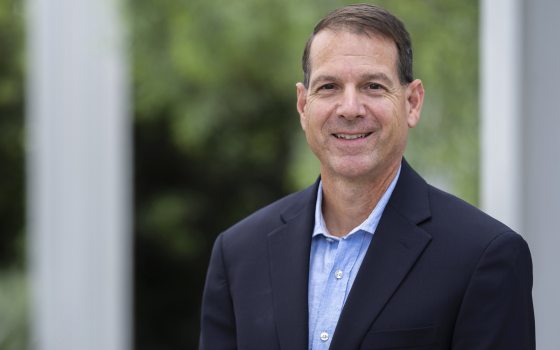 Headshot of a smiling middle-aged man with short dark hair, wearing a navy suit jacket and a light blue collared shirt, with a blurred green garden and white columns in the background.