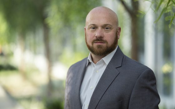 Headshot of a bald man with a full beard, wearing a gray suit jacket and a white patterned shirt, standing outdoors with blurred green trees in the background.