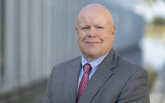Headshot of a bald, red-faced man smiling, wearing a gray suit jacket, light blue shirt, and a magenta patterned tie, standing outdoors in front of a modern gray building.