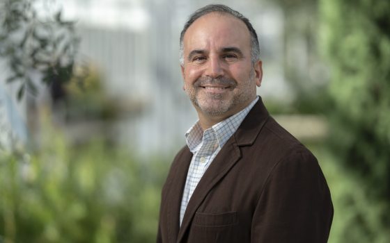 Headshot of a man with short graying hair and a beard, wearing a brown sport coat and a light blue checkered shirt, smiling outdoors with blurred green foliage in the background.