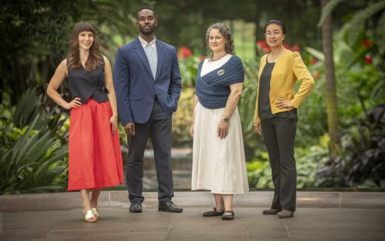 A full-body group portrait of four Longwood Fellows standing side-by-side in formal business-casual attire against a lush green background.