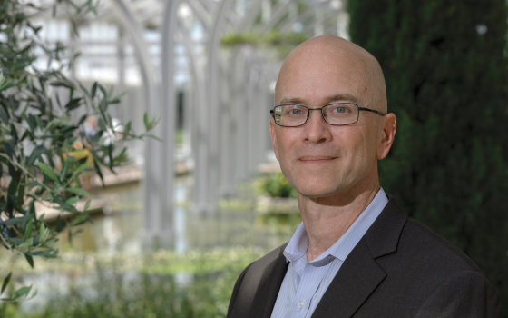 A professional headshot of a smiling man with a shaved head and glasses, wearing a dark blazer over a light blue button-down shirt. He is positioned in the right foreground of the frame. The background features a bright, airy conservatory or greenhouse with tall white architectural arches and greenery reflecting in a calm body of water.