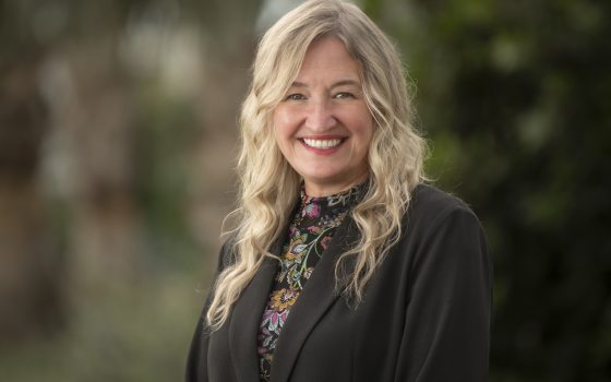 A headshot of a person in a black jacket, with floral shirt, long blond curled hair, smiling at the camera. 