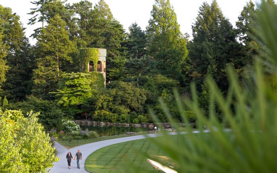 Two guests walk along a curved garden path amid lush greenery and a stone carillon tower in the background.