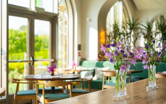 Closeup of pink and purple flowers in two glass vases on a wooden counter, with tables set for guests and large backlit windows in background.