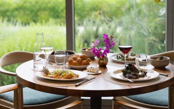 A table in a restuarant with plates of food and beverages, untouched, overlooking a green outdoor space.