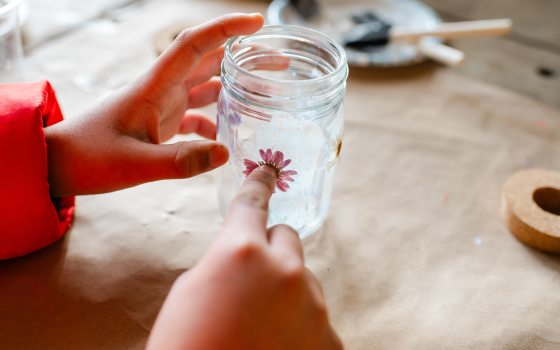 A close-up shot of hands carefully pressing a small, pink dried flower onto the side of a glass jar, showing the intricate detail of the craft project.