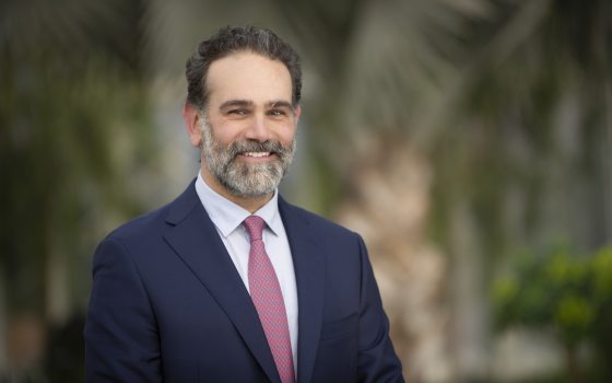 A person in a white shirt, black blazer, and salt and pepper beard smiling at the camera against a blurred garden setting.