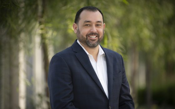A headshot of a person in a white dress shirt and black blazer, against a blurred garden setting.