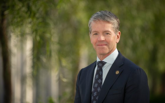 A headshot of a person with salt and pepper hair, in a dark blue coat and tie, slightly smiling at the camera againsta  blurred garden setting.