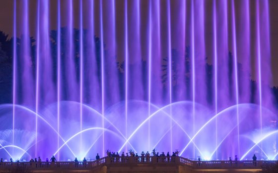 Fans and jets of fountain water bathed in lavender light rise above silhouettes of guests on a stone observation balcony.