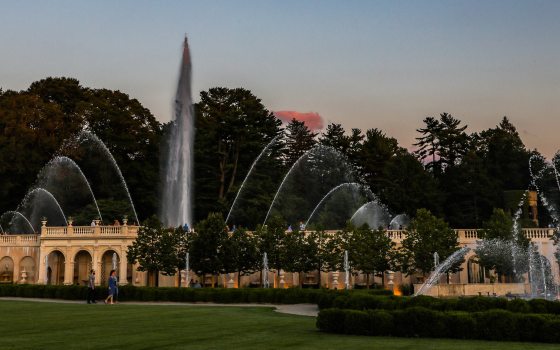 Wide view of a fountain garden at dusk, against the silhouette of trees and a clear deep blue sky.