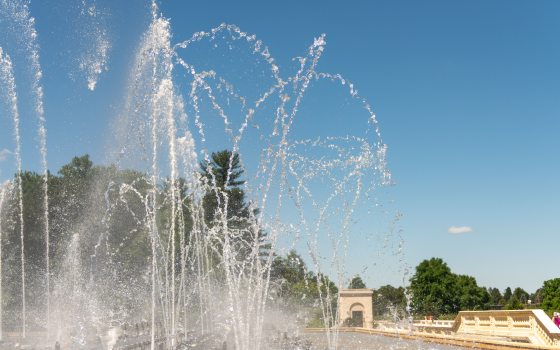 Fountain jets of dancing water rise up from a stone basin against a cloudless blue sky.
