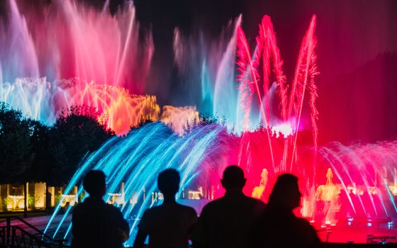 Four foreground silhouettes watch a nighttime fountain show in red, blue, and gold