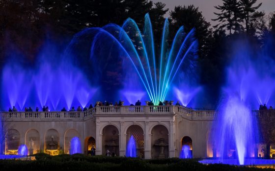 Fountains of water rise in hues of blue from the front and top of a stone facade.
