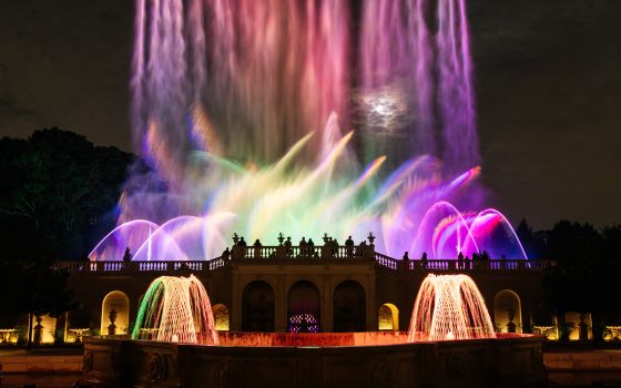 Two basketweave fountains in the foreground frame silhouettes of guests watching pastel-colored fountains fill the night sky in the background/
