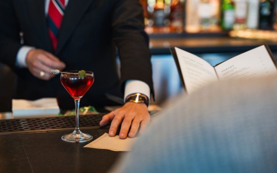 Closeup of a finely dressed bartender presenting a red cocktail with decorative toothpick to a guest seated at the bar
