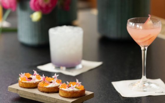 Closeup of two cocktails and a small board with appetizers, with large fuchsia blooms in the background.
