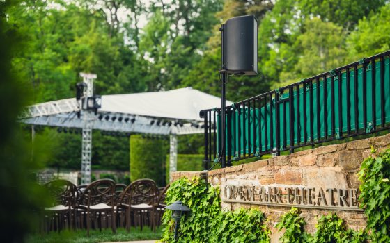 A stone half wall covered in Ivy says "Open Air Theatre." The background depicts a tree-filled setting with a white tent over a large stage with seats on a lawn.  